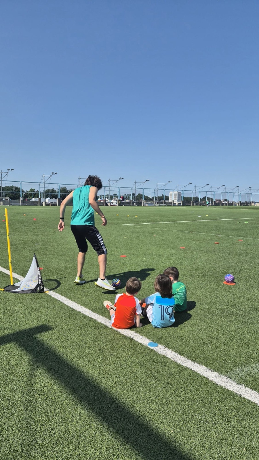 Coach Sam running a drill while three young players sit and watch on the turf field